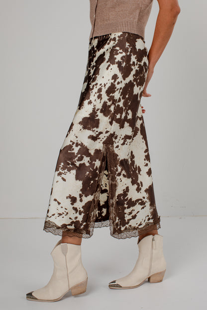 A model is shown from the waist down wearing a brown and cream cow-print skirt with a lace hem, paired with cream ankle boots and a beige knit top, set against a plain light gray studio background.