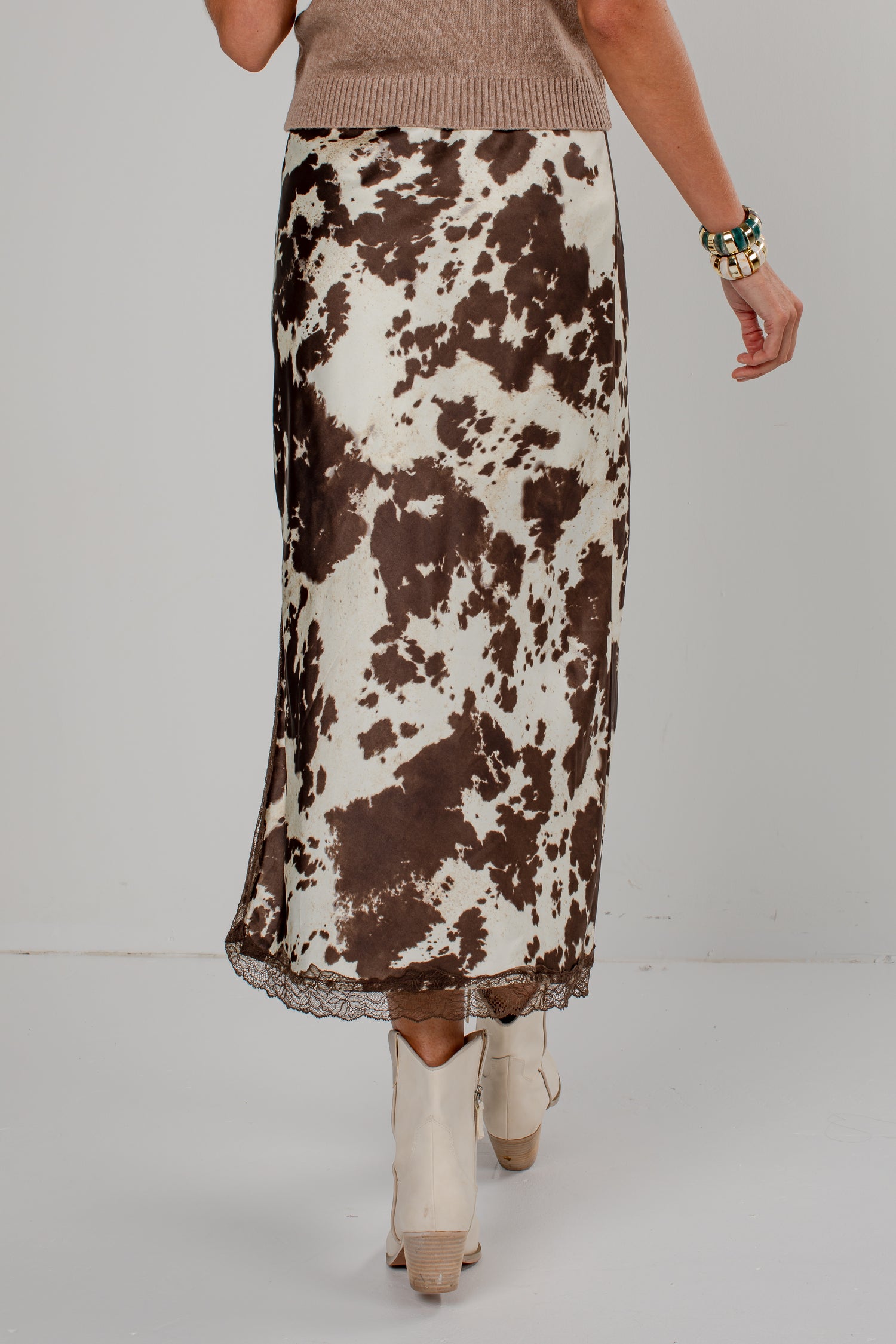 A model is shown from the waist down wearing a brown and white cow-print midi skirt with a lace hem, paired with cream-colored pointed-toe ankle boots.