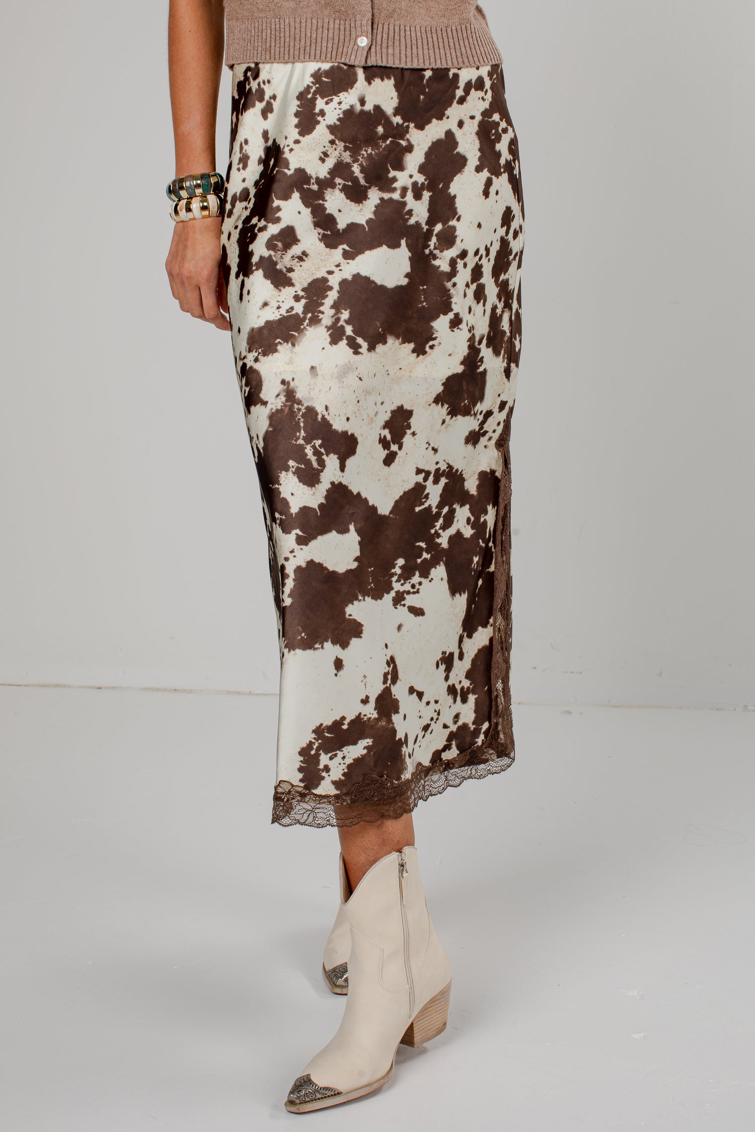 A model is shown from the waist down wearing a brown and white cow-print midi skirt with a lace hem, paired with cream-colored pointed-toe ankle boots.