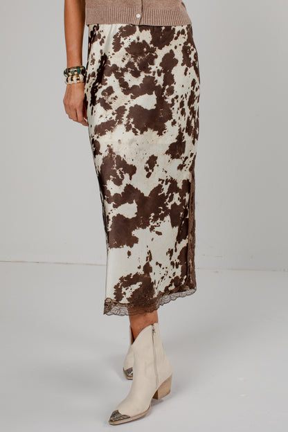 A model is shown from the waist down wearing a brown and white cow-print midi skirt with a lace hem, paired with cream-colored pointed-toe ankle boots.