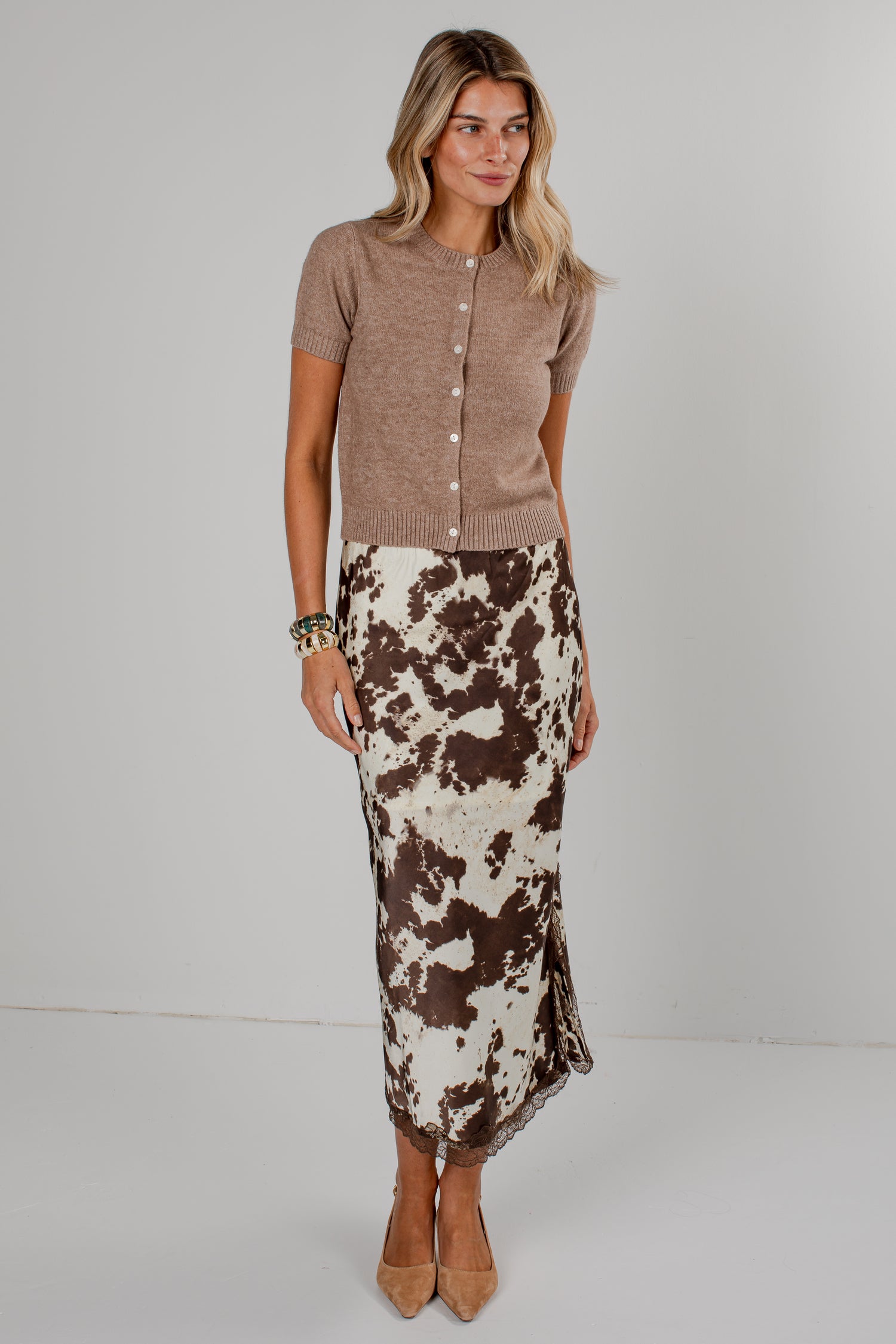 A woman modeling a short-sleeve taupe knit cardigan paired with a brown and cream cow-print midi skirt, standing against a light gray studio background.