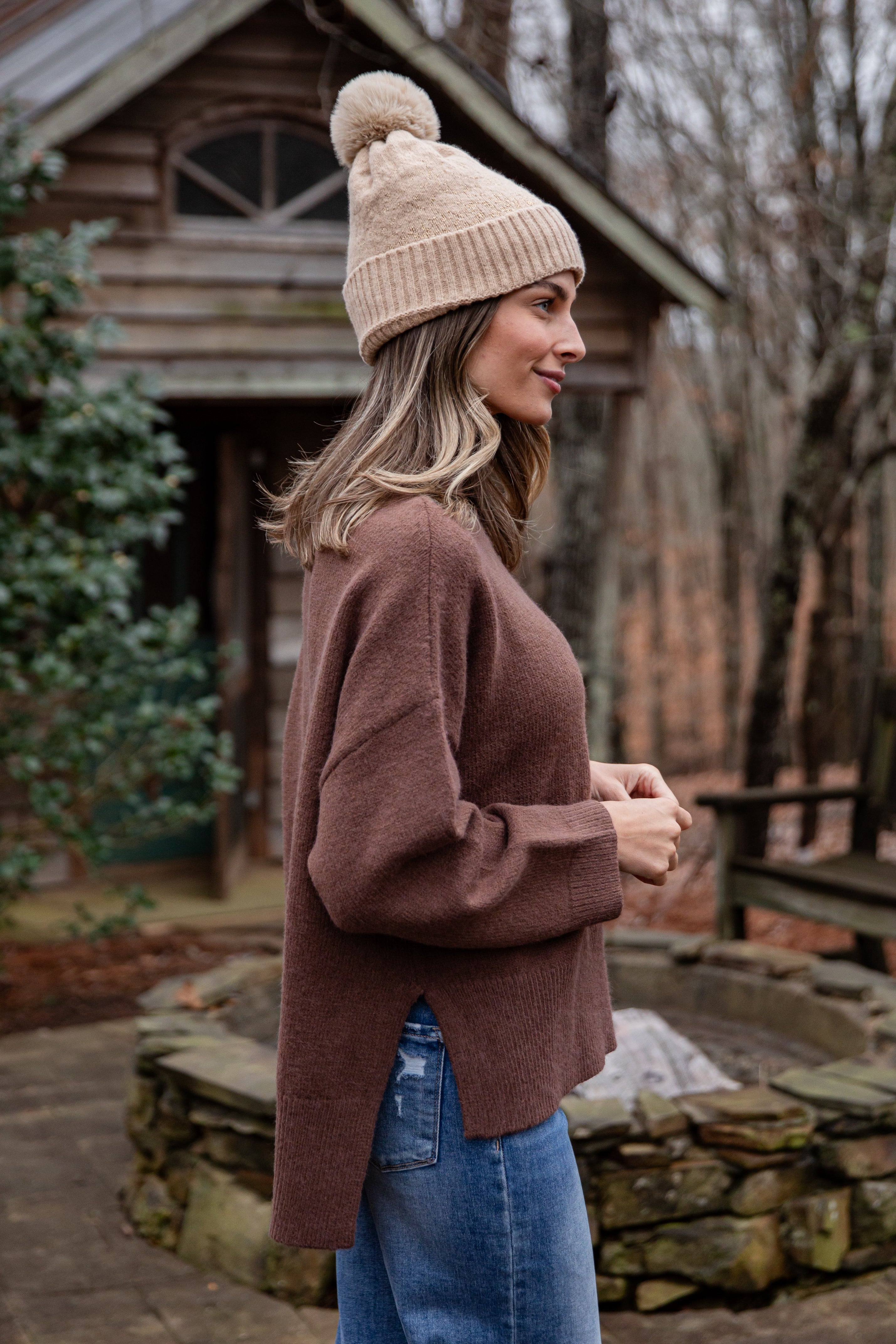 Woman wearing a brown sweater and beige beanie standing outdoors near a wooden cabin.