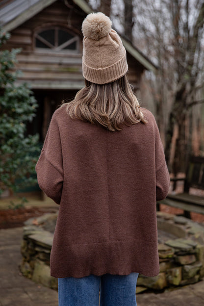 Person wearing a brown coat and beige beanie with pom-pom, standing in front of a wooden cabin.
