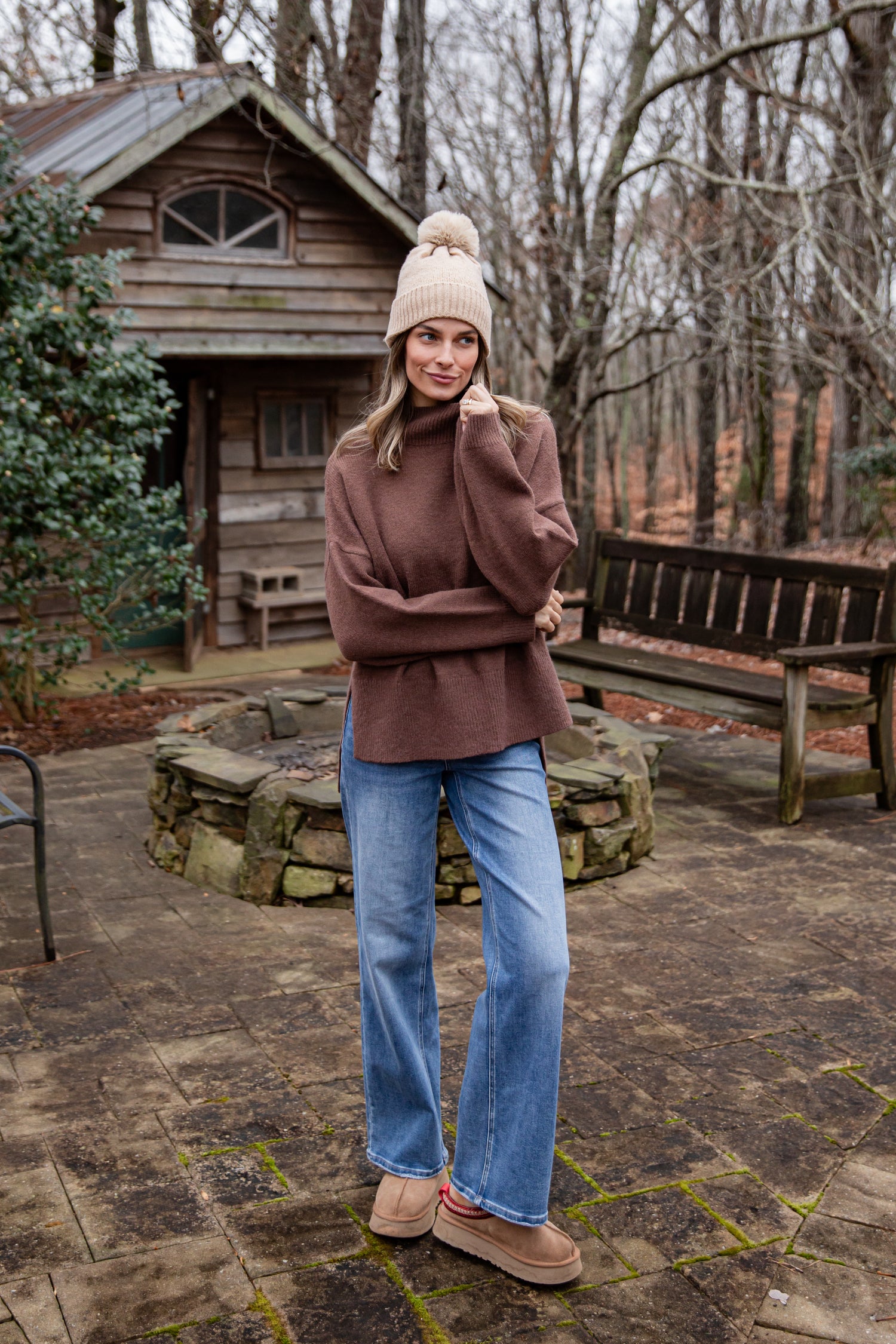 Woman in a brown sweater and jeans standing in front of a wooden cabin.
