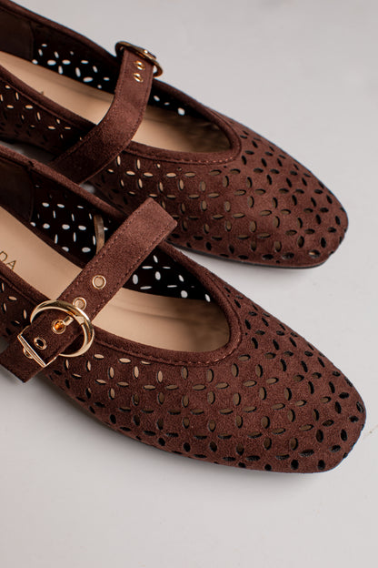 A pair of dark brown suede Mary Jane flats with floral cutout detailing, featuring a single strap with a gold-tone buckle, shown on a light gray background.