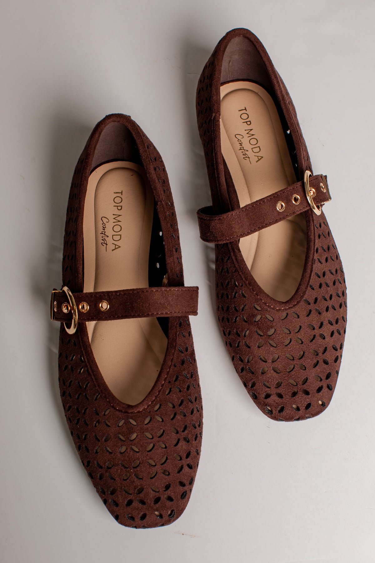 A pair of brown perforated Mary Jane flats with gold-tone buckle straps, shown from a top-down angle on a light gray background. The brand name &