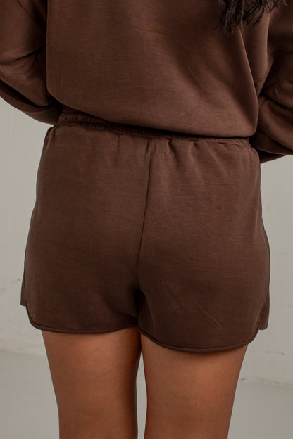 A close-up back view of a person wearing matching brown lounge set, featuring a long-sleeve top and coordinating shorts with an elastic waistband.