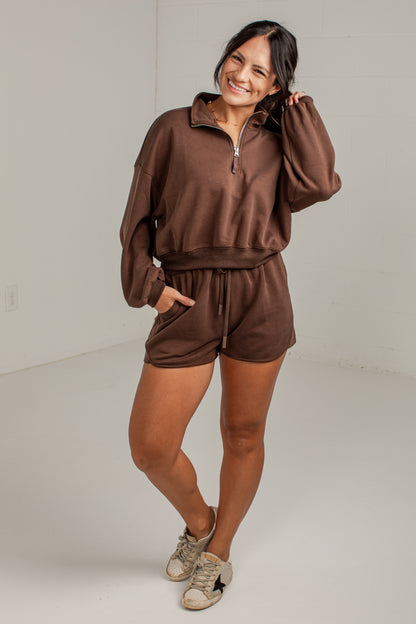 A woman modeling a matching brown lounge set consisting of a half-zip sweatshirt and matching shorts, paired with beige sneakers, in a bright studio setting.