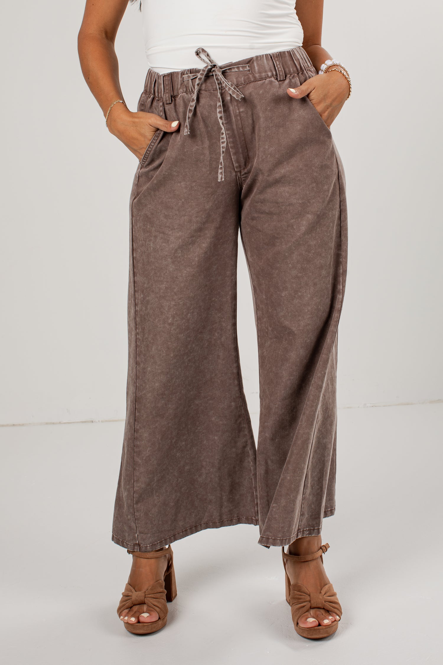 A person wearing high-waisted, wide-leg brown pants with a drawstring waist, paired with a white top and brown heeled sandals, photographed against a plain light gray studio background.