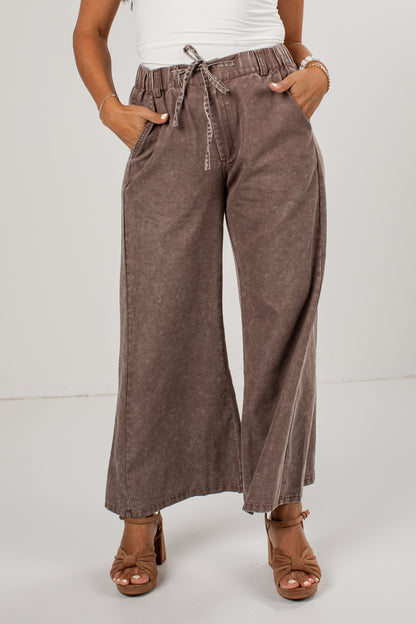 A person wearing high-waisted, wide-leg brown pants with a drawstring waist, paired with a white top and brown heeled sandals, photographed against a plain light gray studio background.
