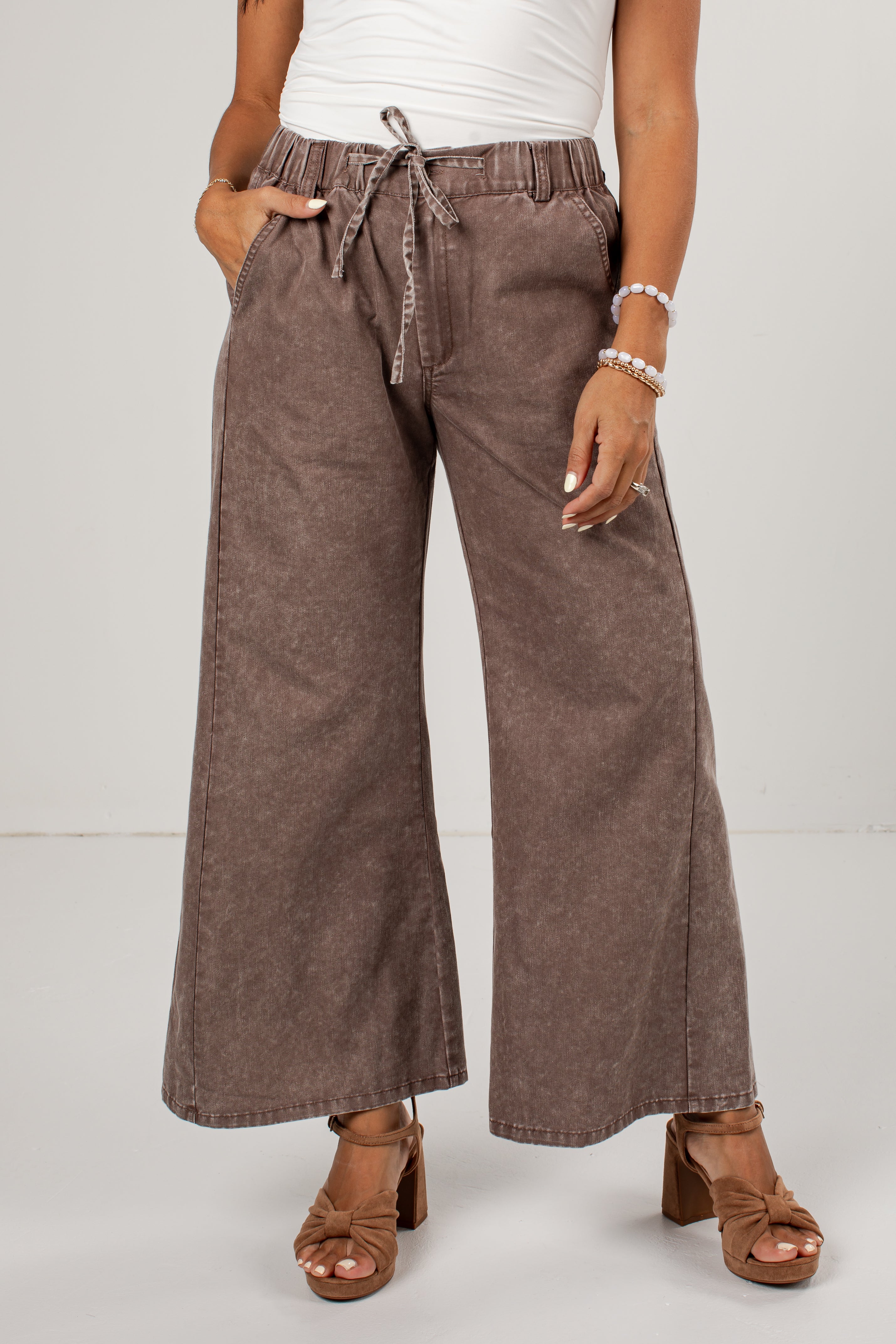 A person wearing wide-leg cropped pants in a washed taupe-brown color, styled with a white top and brown block-heel sandals against a light gray studio background.