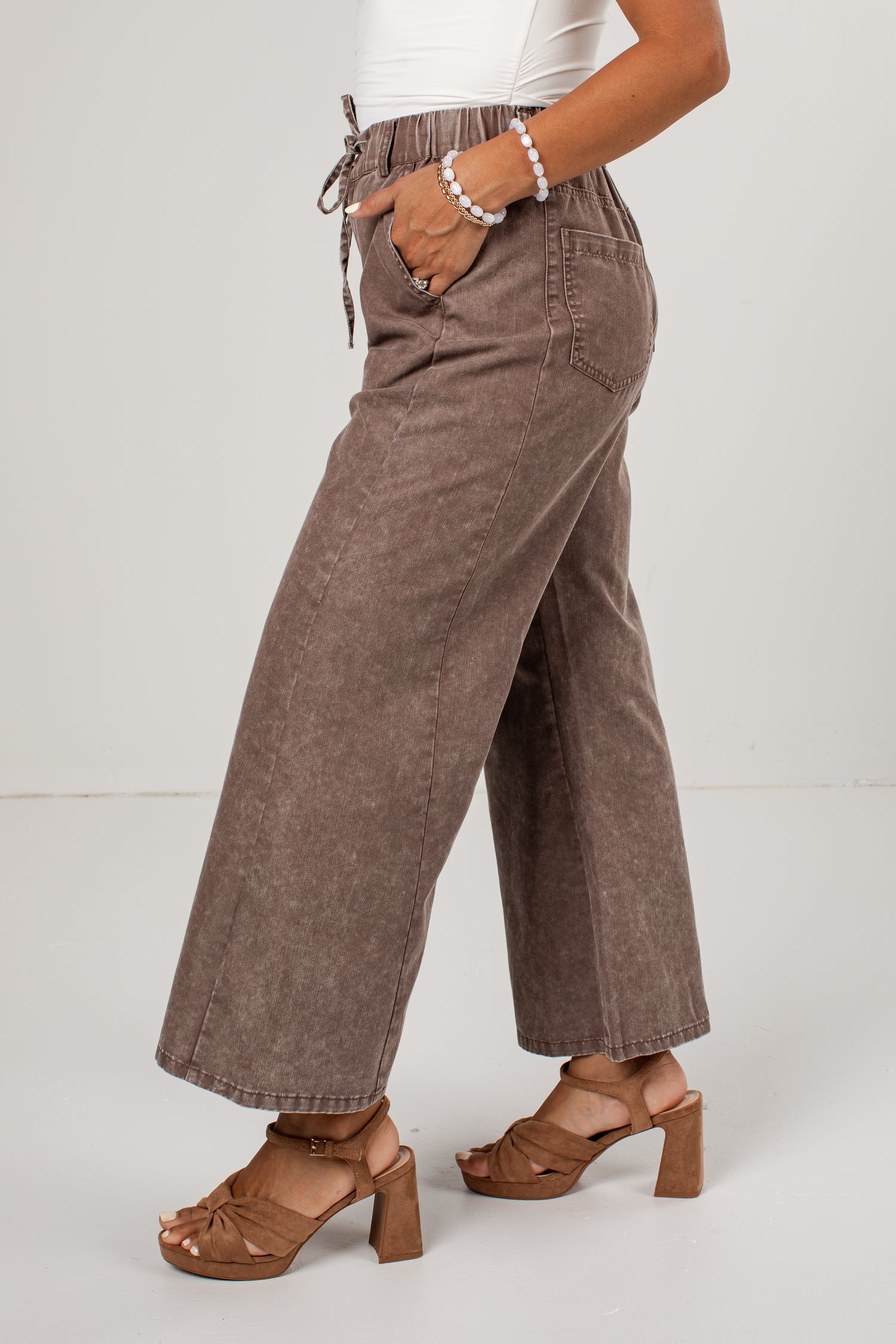 A person wearing wide-leg brown corduroy pants with a drawstring waist, paired with brown heeled sandals and a white top, shown from the waist down against a light gray studio background.