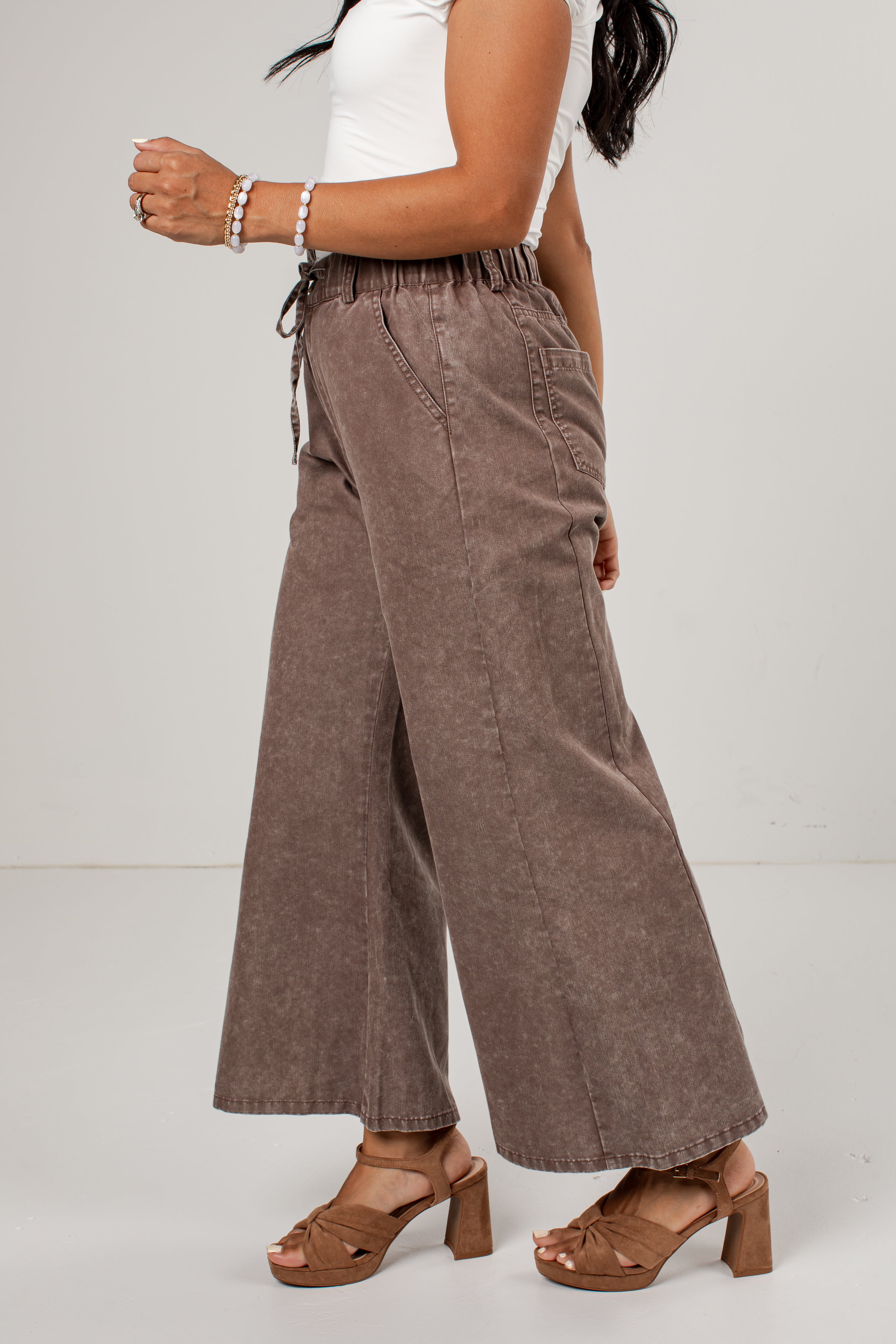 A model wearing high-waisted, wide-leg brown corduroy pants with an elastic waistband and drawstring, paired with a white tank top and brown block-heel sandals against a light gray studio background.