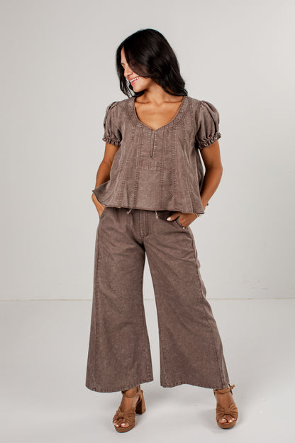 A woman modeling a matching two-piece outfit consisting of a short-sleeve peplum top and wide-leg pants in a textured taupe-brown fabric, paired with brown heeled sandals against a plain light gray studio background.