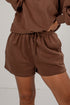 A close-up of a person wearing brown drawstring shorts and a matching brown sweatshirt against a plain light background.