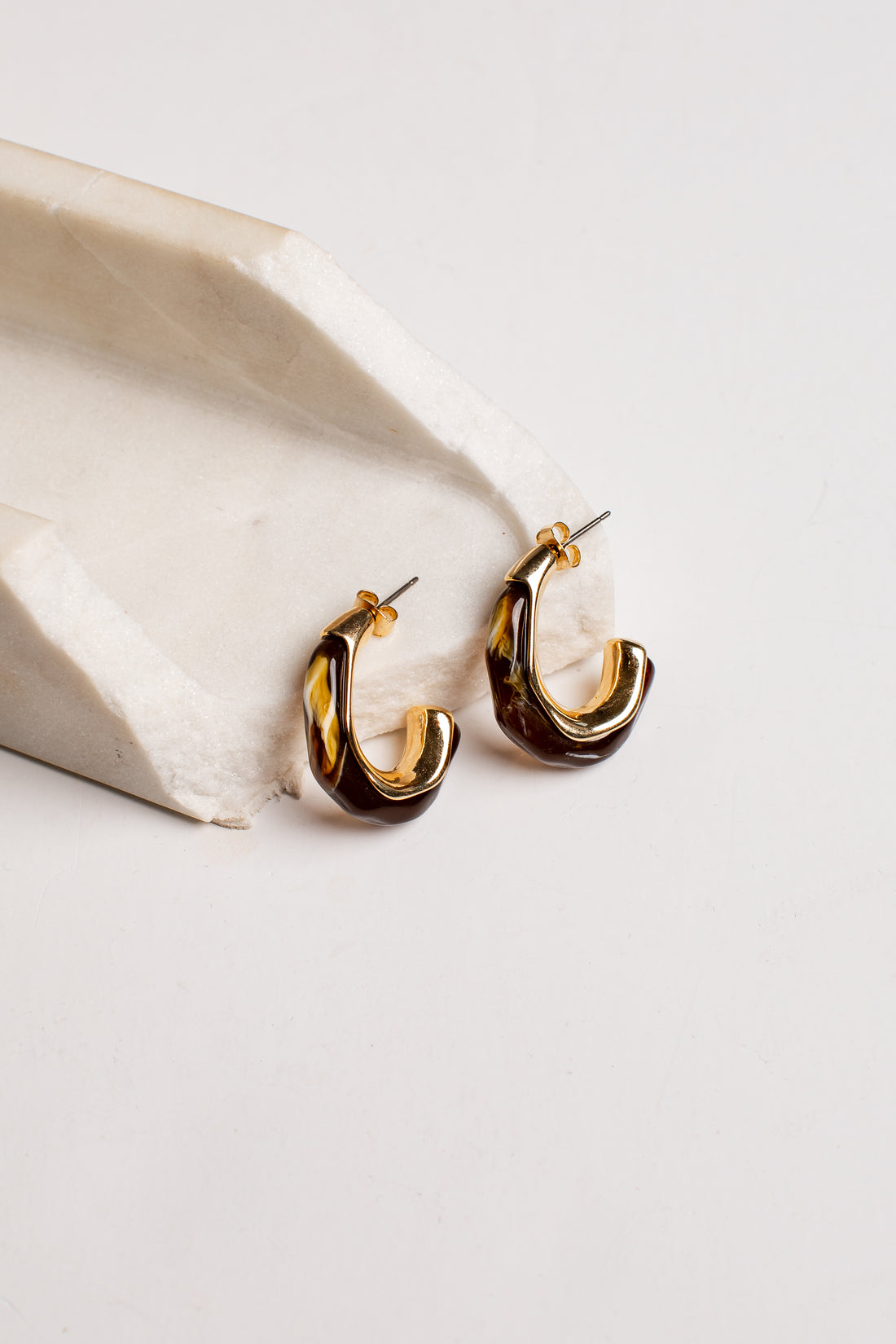 A pair of gold and dark brown twisted hoop earrings resting on a light beige stone prop against a clean white background.