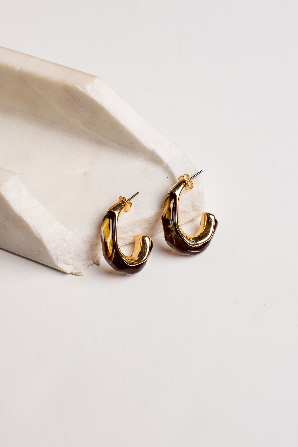 A pair of gold and dark brown twisted hoop earrings resting on a light beige stone prop against a clean white background.
