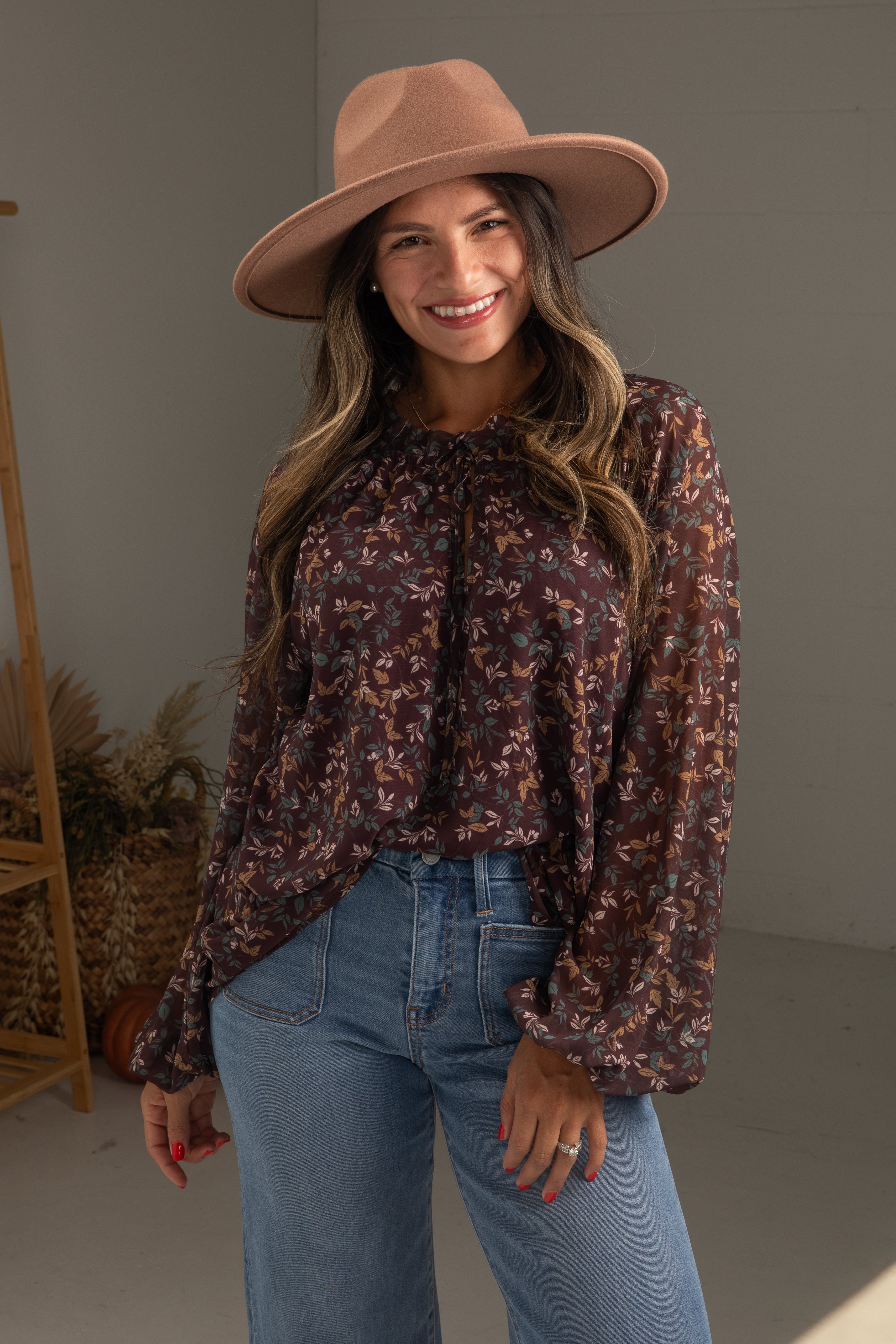 Woman wearing a floral blouse, jeans, and a wide-brimmed hat indoors.