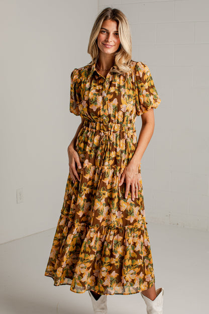 A woman modeling a short-sleeve midi dress with a yellow, orange, and brown floral pattern, paired with white cowboy boots, standing against a white brick wall.
