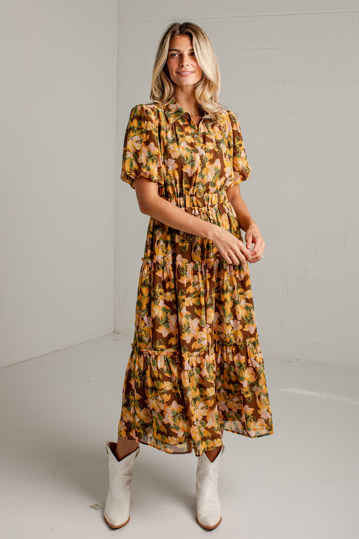 A woman modeling a short-sleeve, tiered midi dress with a warm-toned floral print in yellow, orange, and brown, paired with white cowboy boots against a white studio background.