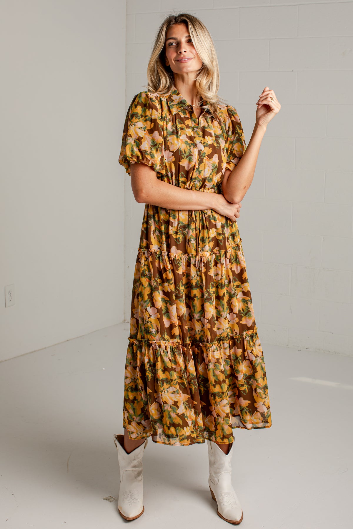 A woman modeling a short-sleeve tiered midi dress with a yellow, orange, and green floral pattern, paired with white cowboy boots, standing against a white studio wall.