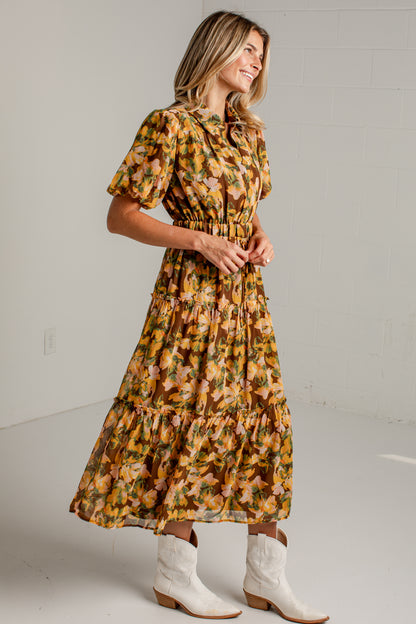 A woman modeling a short-sleeve, tiered midi dress with a yellow, orange, and brown floral pattern, paired with white cowboy boots, standing against a white studio wall.