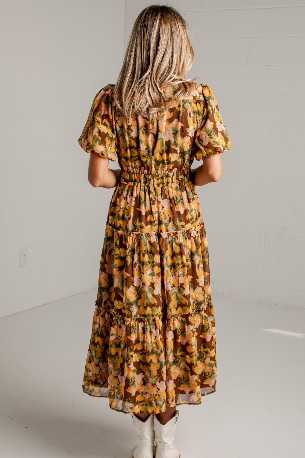 A woman viewed from behind wearing a tiered midi dress with puff sleeves and an allover floral print in mustard yellow, orange, and green, paired with white cowboy boots in a bright studio setting.