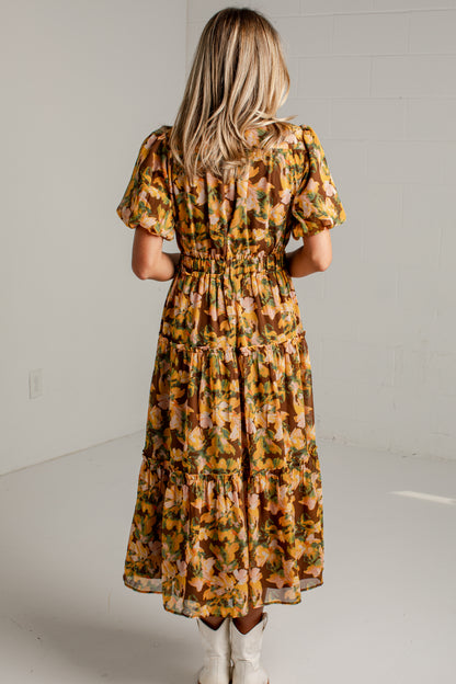 A woman viewed from behind wearing a tiered midi dress with puff sleeves and an allover floral print in mustard yellow, orange, and green, paired with white cowboy boots in a bright studio setting.