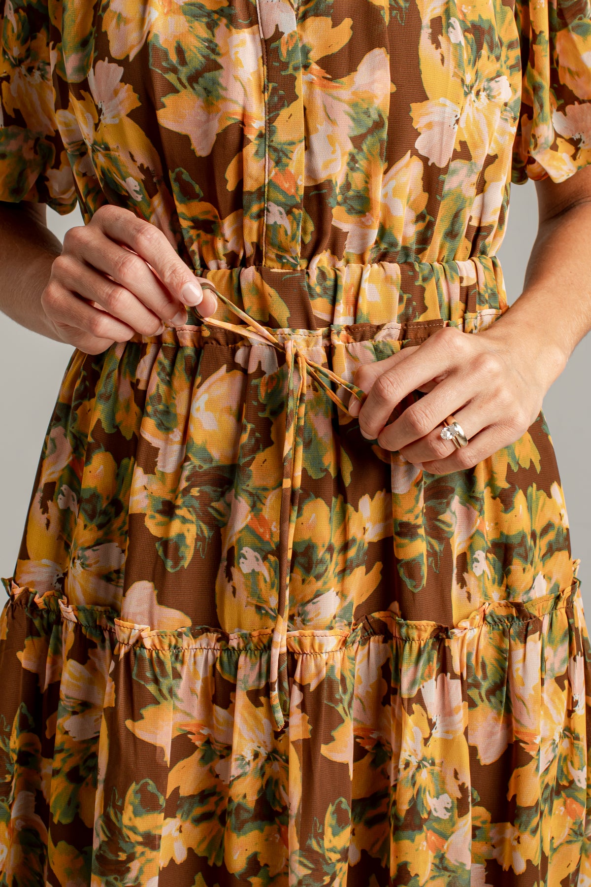 Close-up of a woman wearing a brown tiered dress with a yellow, orange, and green floral print, tying the drawstring belt at her waist.