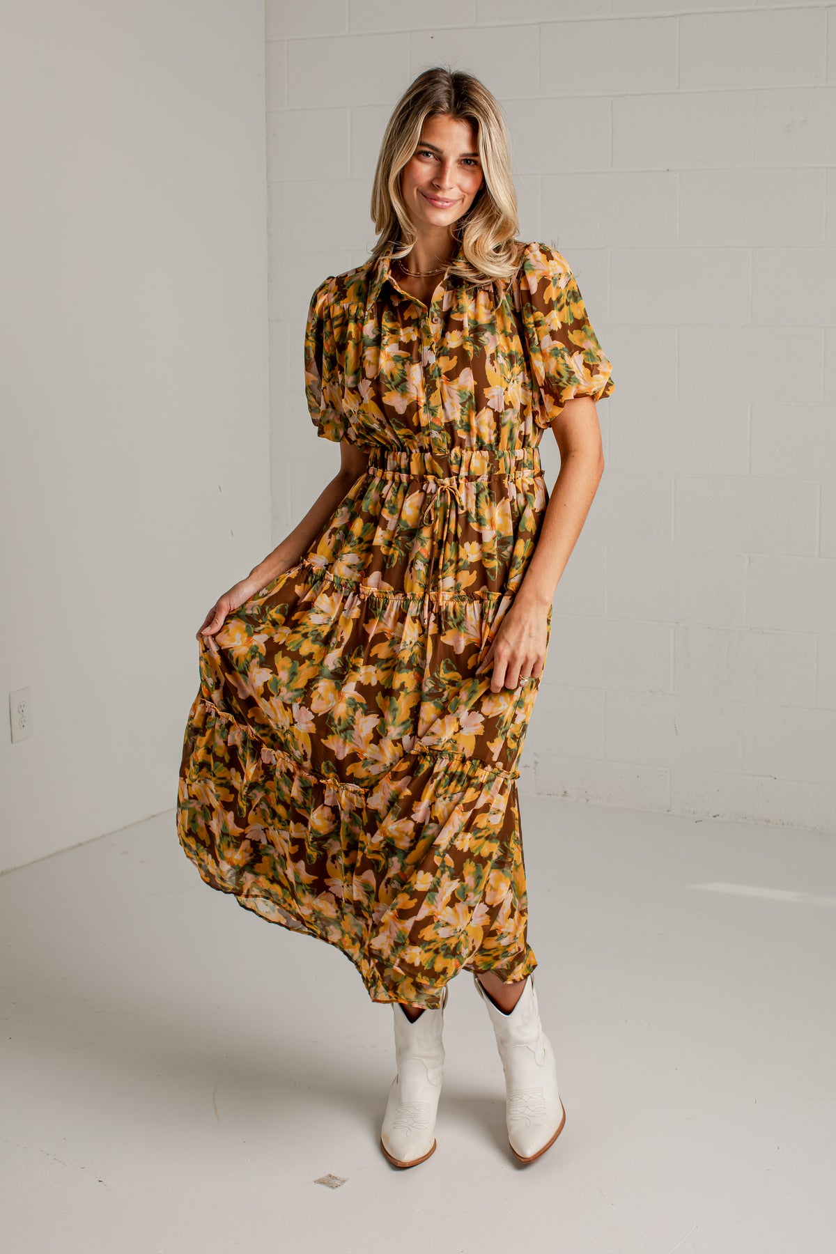 A woman modeling a short-sleeve midi dress with a yellow, orange, and green floral pattern, paired with white cowboy boots, standing against a white brick wall.