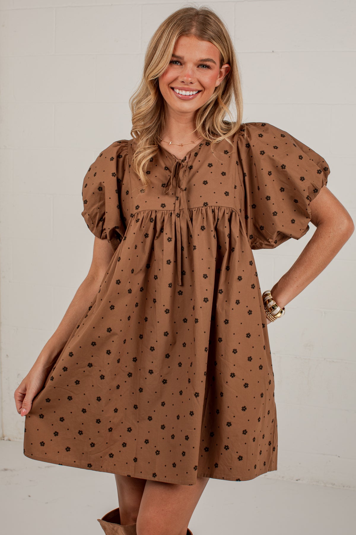 A woman modeling a brown short-sleeve dress with black polka dots, puff sleeves, and a tie neckline, set against a white brick wall.
