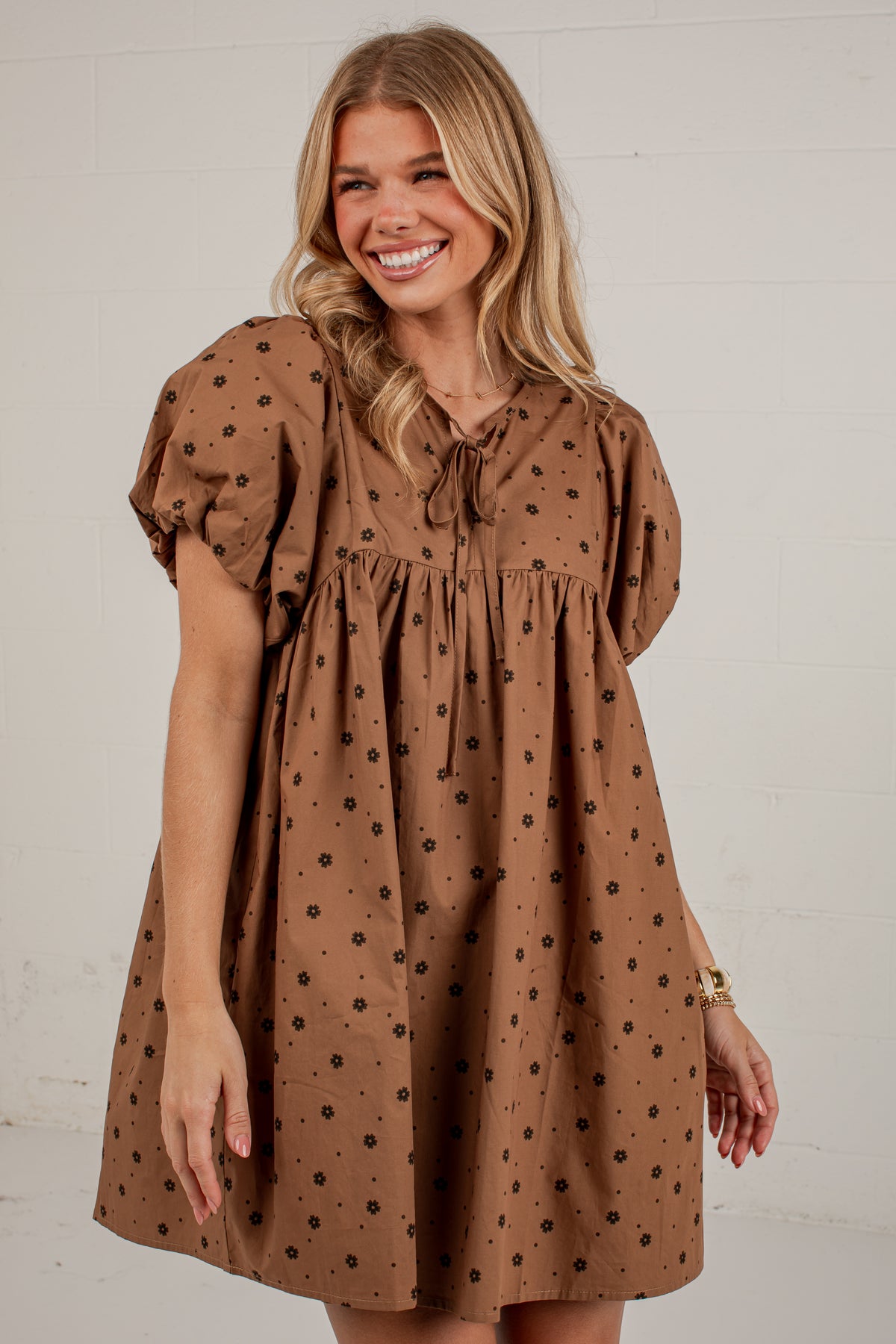 A smiling blonde woman wearing a brown short-sleeve dress with black flower dots, puff sleeves, and a tie neckline, standing against a white brick wall.