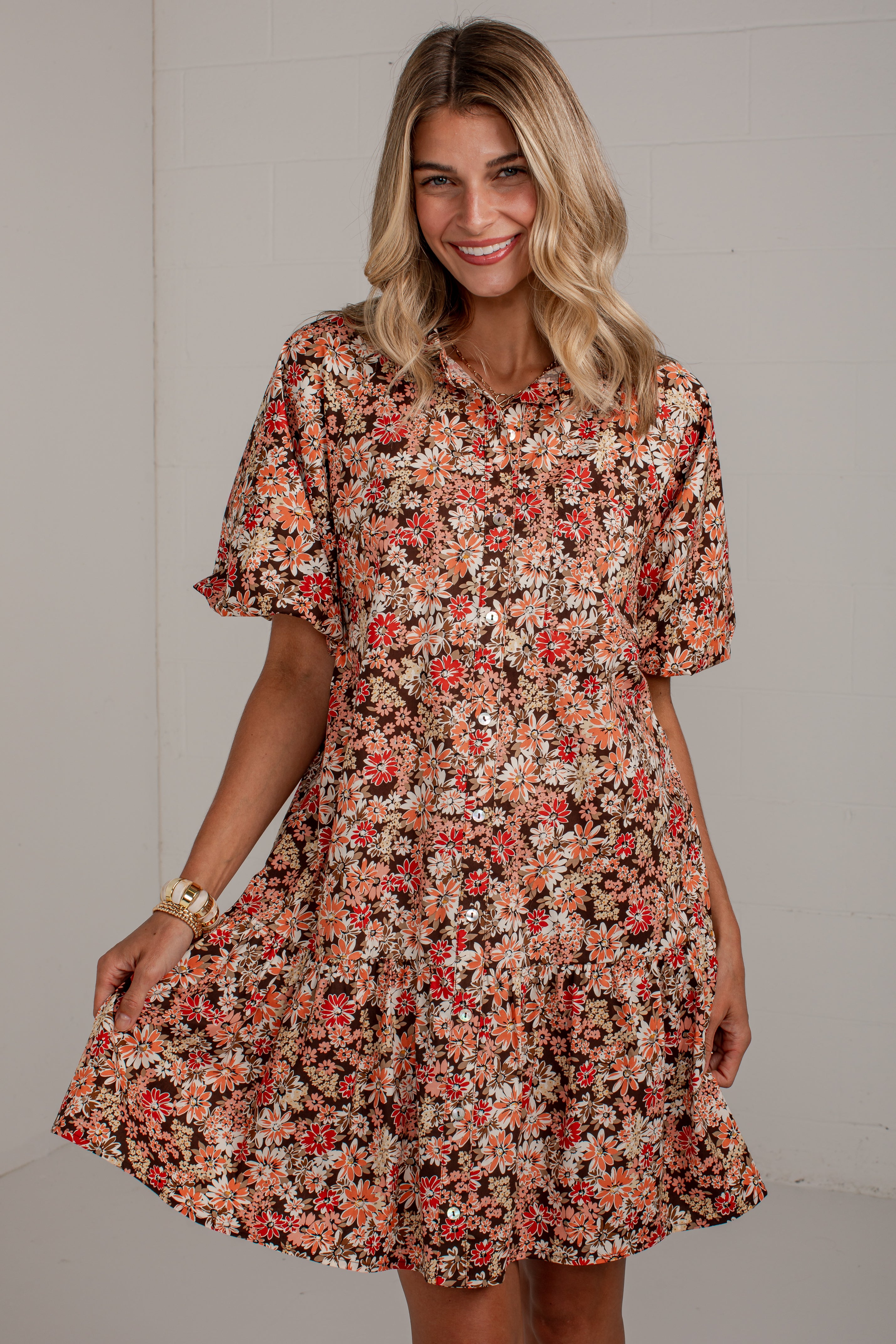 A woman modeling a short-sleeve floral dress with a button-up front and flared skirt, featuring an all-over print in orange, red, cream, and brown tones against a light gray wall.