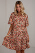 A woman modeling a short-sleeve floral dress with a button-up front and flared skirt, featuring an all-over print in orange, red, cream, and brown tones against a light gray wall.