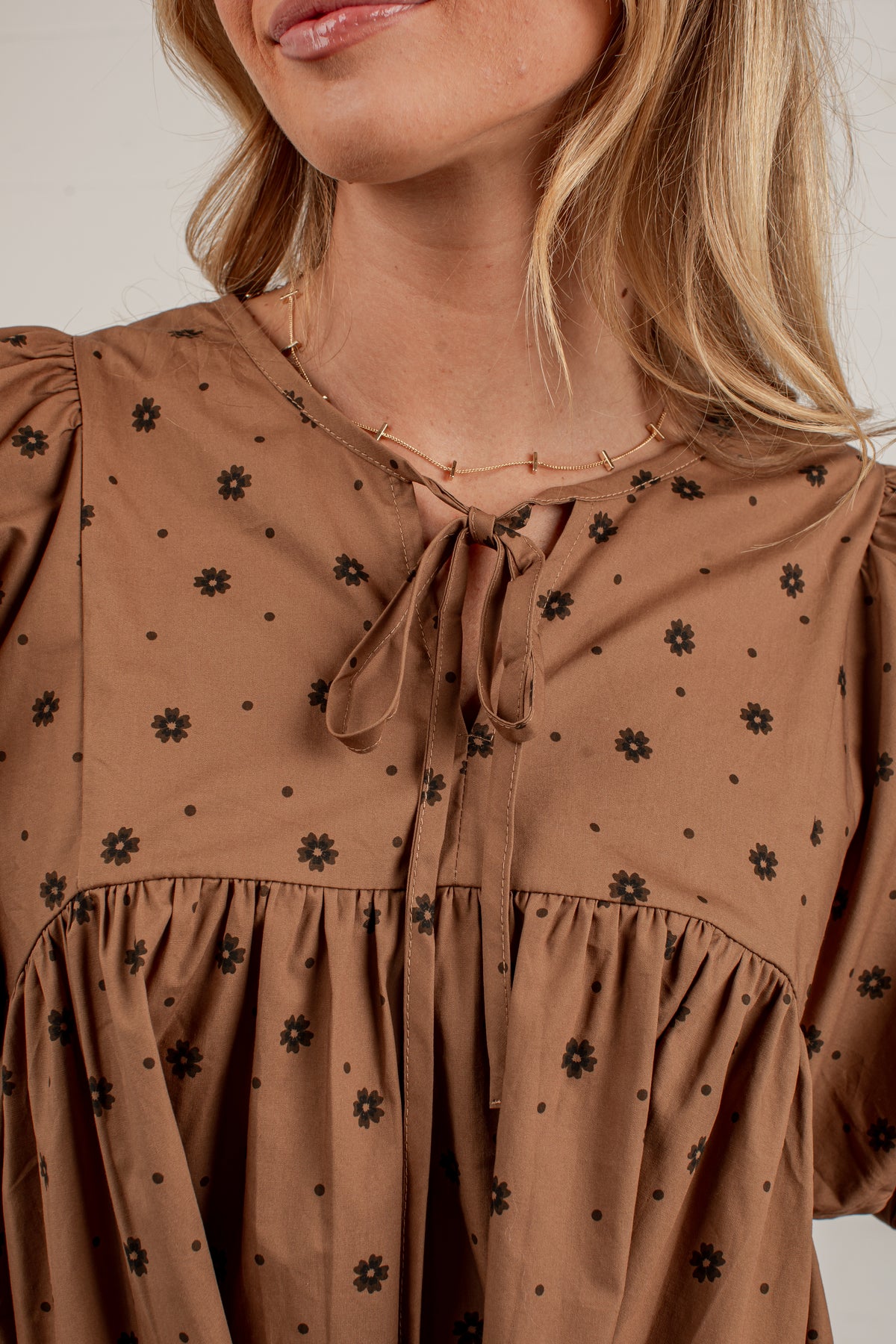 A close-up of a woman wearing a brown blouse with a black floral and polka dot pattern, featuring a tie-neck detail and ruffled sleeves.