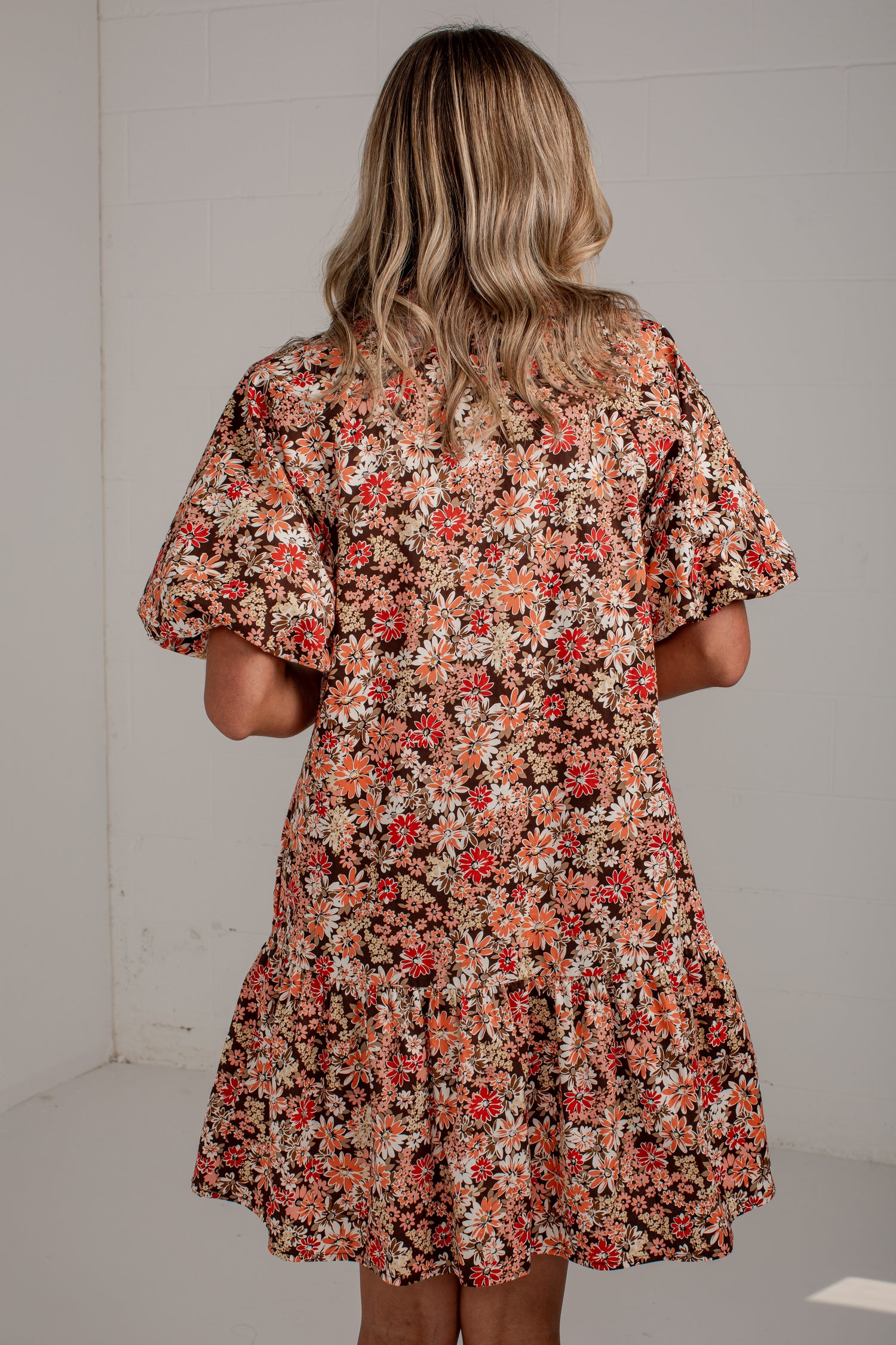 A woman viewed from behind wearing a short-sleeve floral dress with puff sleeves and a tiered skirt, featuring a dense print of red, orange, and white flowers on a dark background.