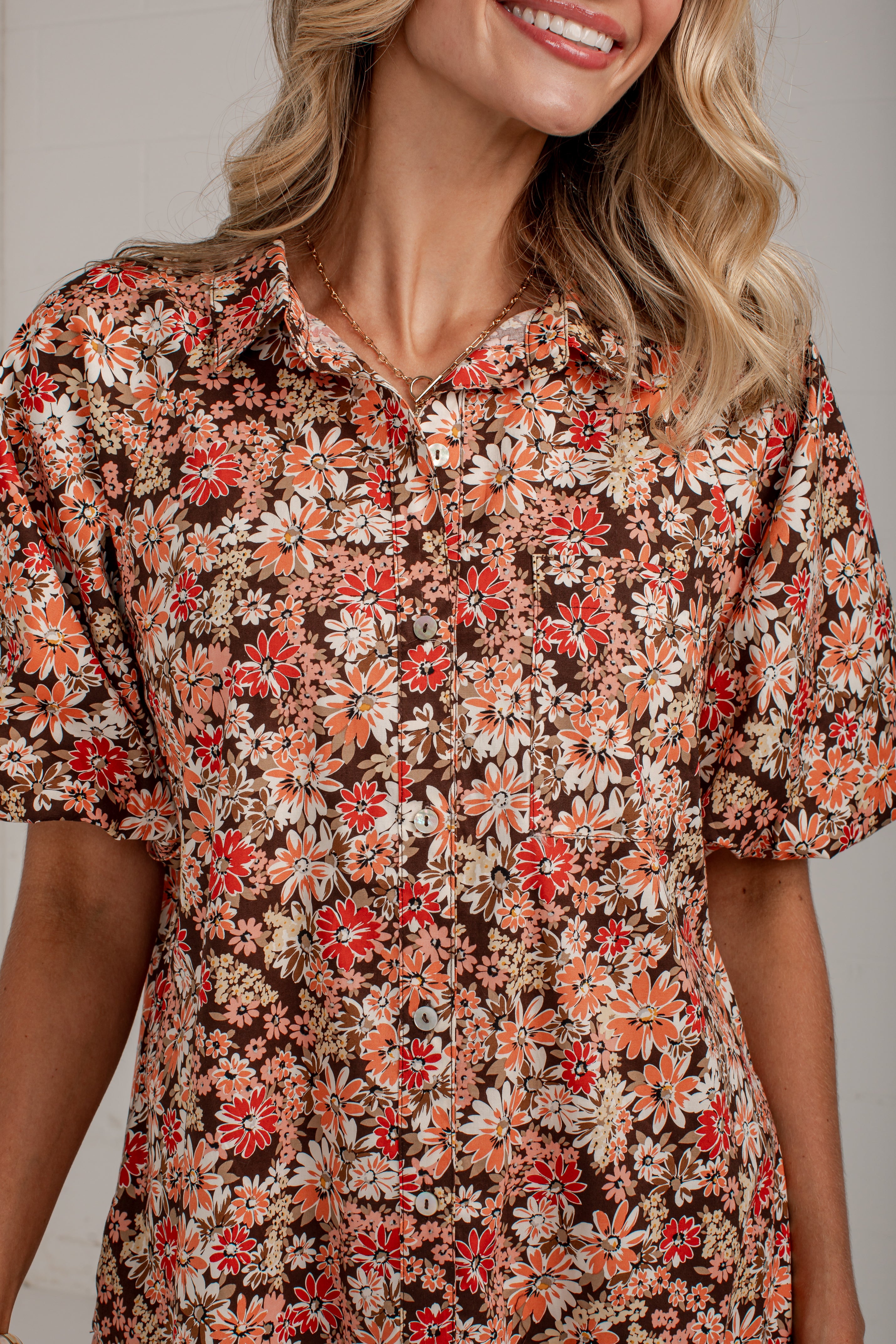 A close-up of a woman wearing a short-sleeve button-front blouse with a dense floral print in red, orange, cream, and white over a dark brown background.