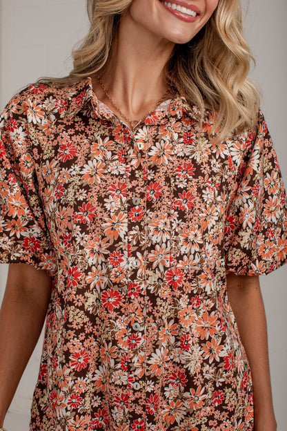 A close-up of a woman wearing a short-sleeve button-front blouse with a dense floral print in red, orange, cream, and white over a dark brown background.