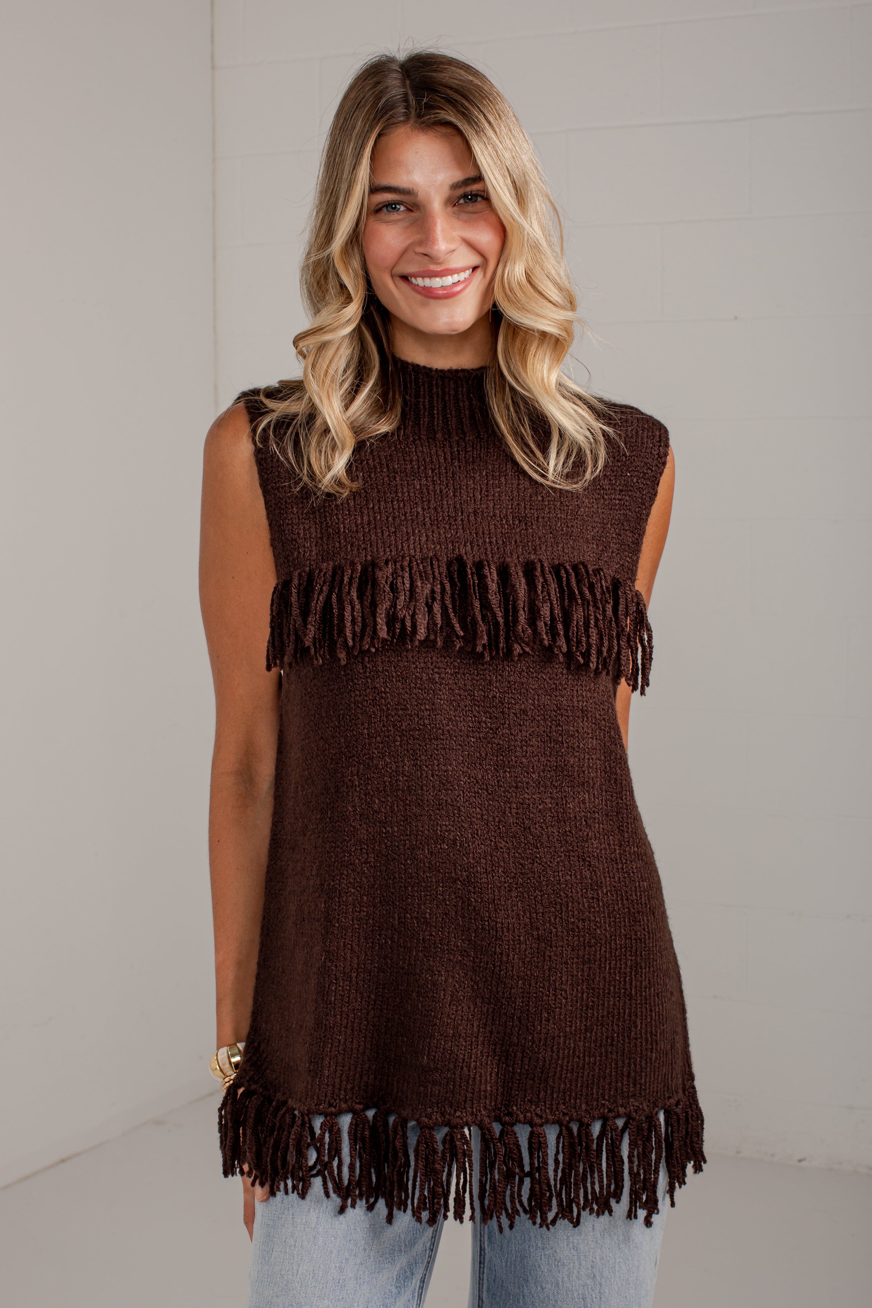 A woman wearing a sleeveless dark brown knit tunic top with fringe trim at the chest and hem, paired with light blue jeans, standing against a light gray wall.