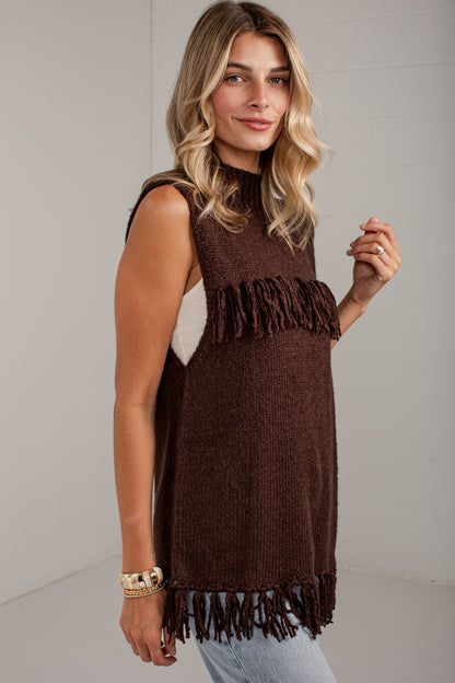 A woman modeling a sleeveless brown knit tunic top with fringe trim, paired with light blue jeans, against a plain light gray studio background.
