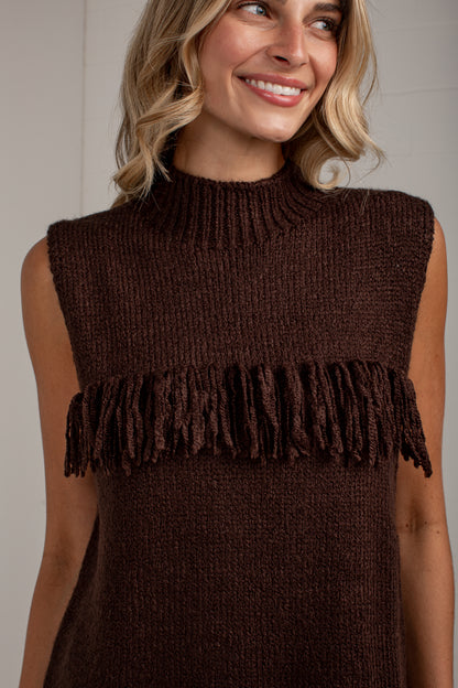 A woman wearing a sleeveless brown knit sweater with a high ribbed mock neck and a fringe detail across the chest, shown against a neutral indoor background.