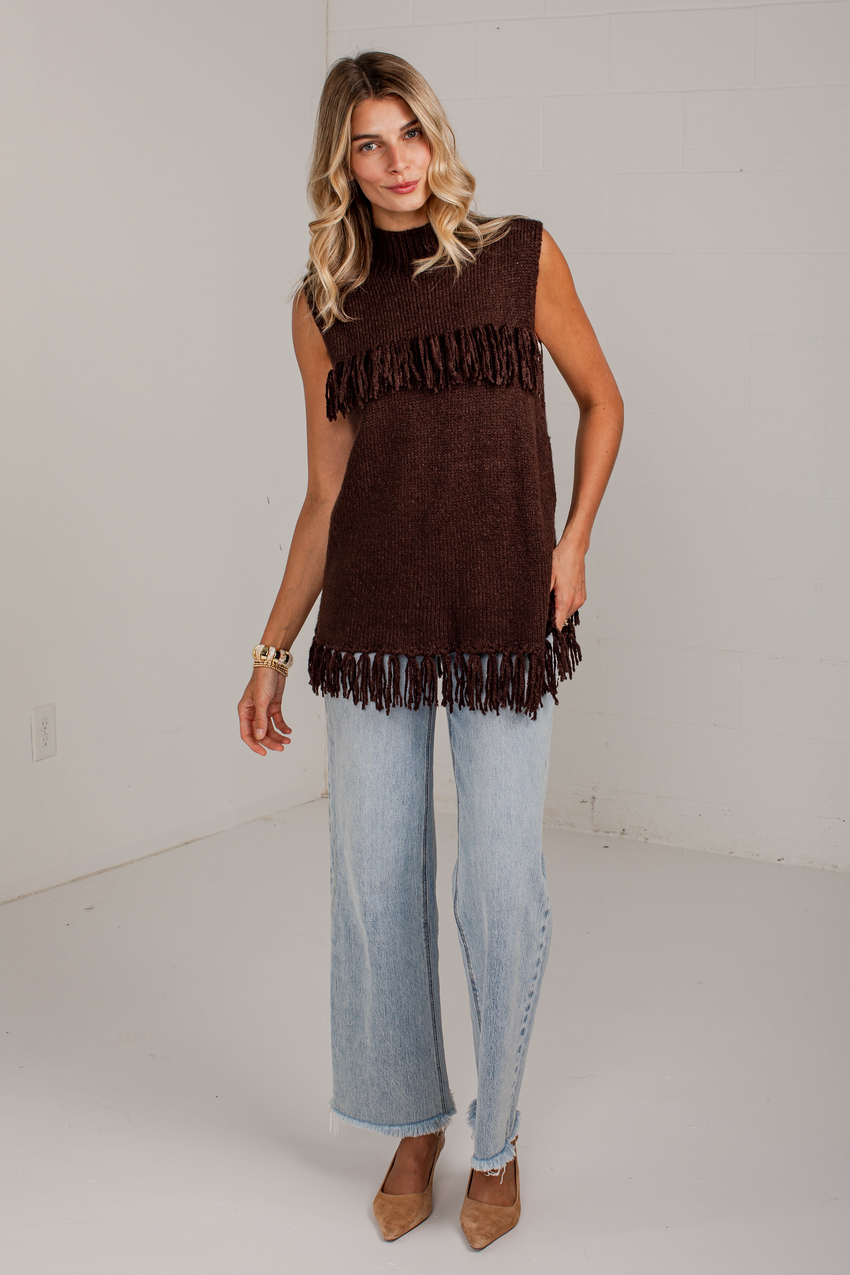 A woman modeling a sleeveless dark brown knit top with fringe trim, paired with light-wash wide-leg jeans and tan pointed-toe heels, standing against a plain white studio wall.