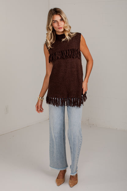 A woman modeling a sleeveless dark brown knit top with fringe trim, paired with light-wash wide-leg jeans and tan pointed-toe heels, standing against a plain white studio wall.