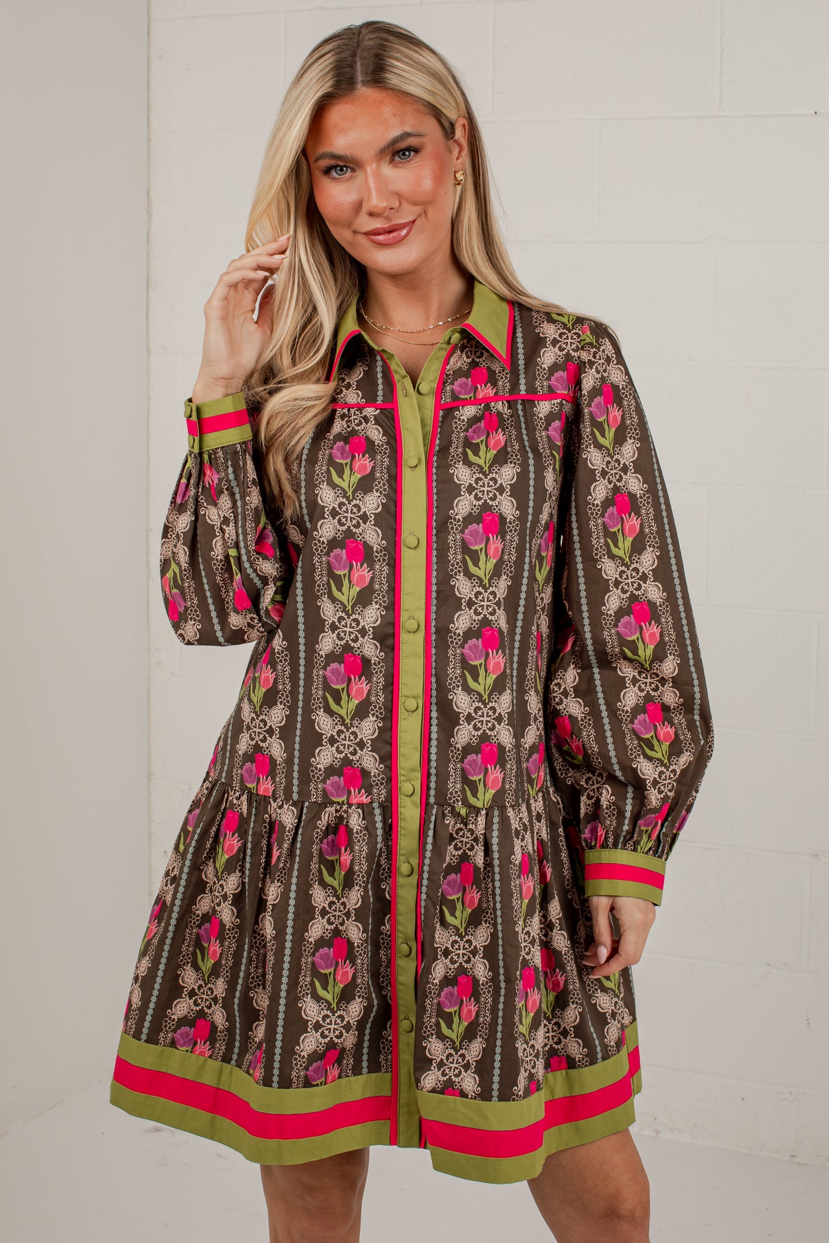 A woman models a brown long-sleeve mini shirt dress with a pink and green floral pattern, featuring a green collar, pink and green trim, and a button-up front.