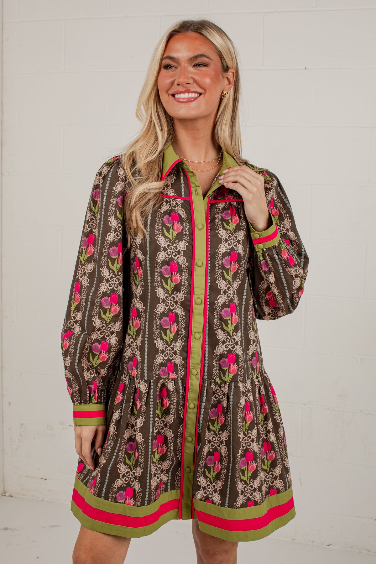 A woman modeling a brown patterned button-up shirt dress with pink floral motifs, green trim, and a green collar, standing against a white brick wall.