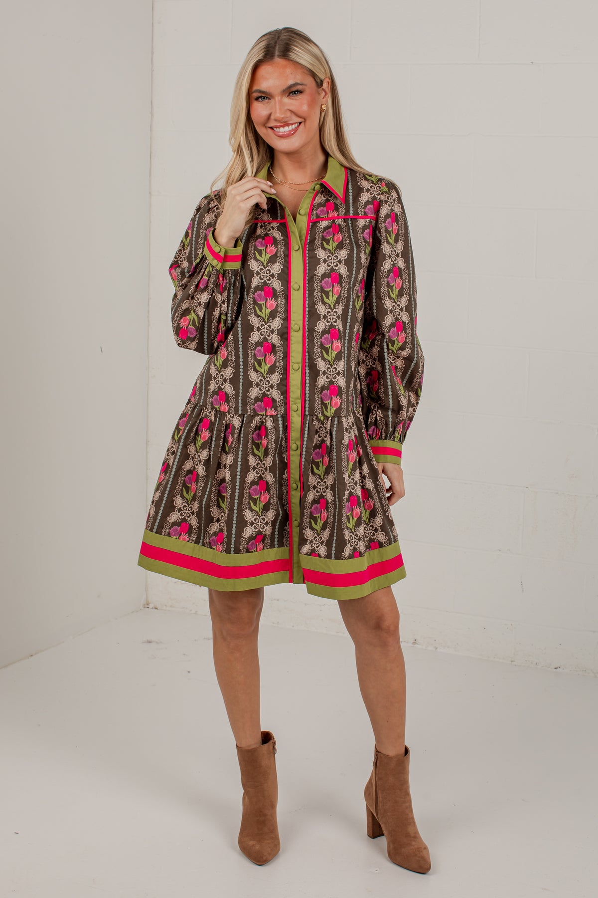A woman modeling a brown patterned long-sleeve shirt dress with pink and green accents, paired with brown ankle boots, in a bright studio setting.