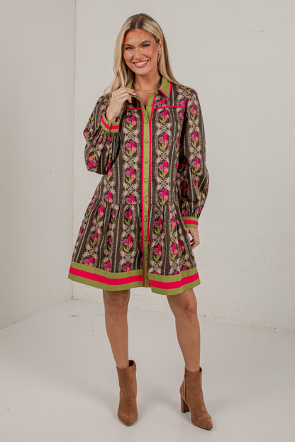 A woman modeling a brown patterned long-sleeve shirt dress with pink and green accents, paired with brown ankle boots, in a bright studio setting.