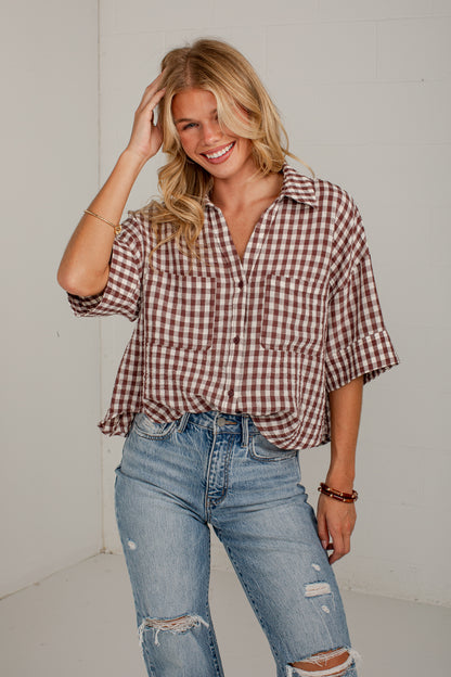 A woman modeling a brown and white gingham short-sleeve button-up shirt with rolled cuffs, paired with distressed blue jeans, standing against a white wall.