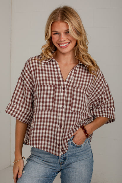 A smiling blonde woman wearing a brown and white gingham check short-sleeve button-up shirt with rolled cuffs and light blue jeans, standing against a light gray wall.