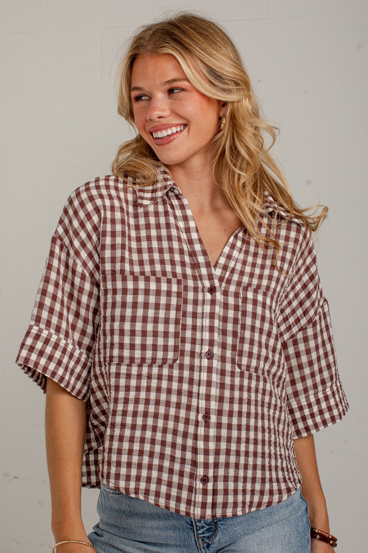 A smiling blonde woman wearing a brown and white gingham check short-sleeve button-up shirt with rolled cuffs and blue jeans, standing against a light gray wall.